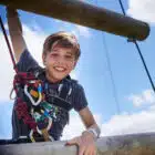 A smiling boy at Multi-Activity, Newby Wiske Hall - Jul 27, 2024 climbs a high rope obstacle under a blue sky with clouds.