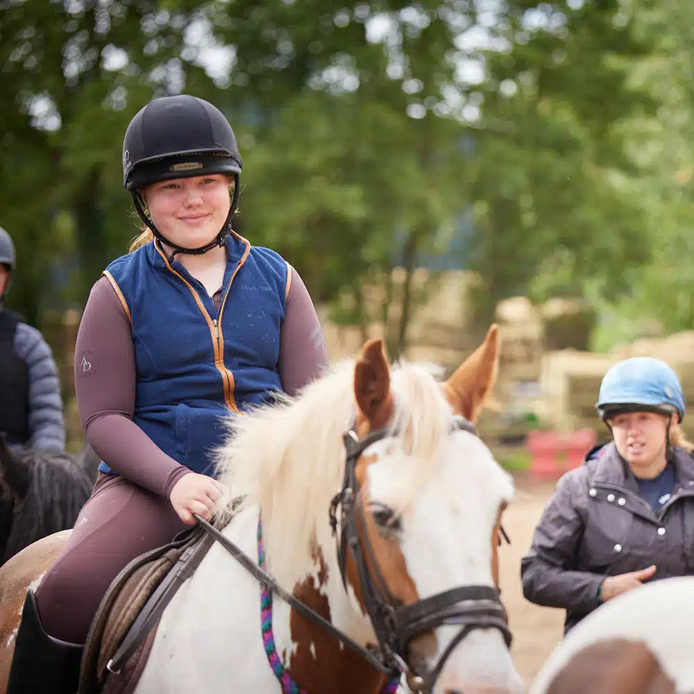 A smiling young rider in a helmet seated on a Pony Camp Trekker, Tregoyd House - Jul 27, 2024, with another rider and horse visible in the background.