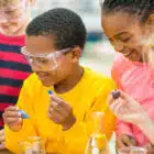 Three children in safety goggles conduct a Streetdance experiment with test tubes and liquids, laughing and engaged in their activity at Boreatton Park on Aug 03, 2024.