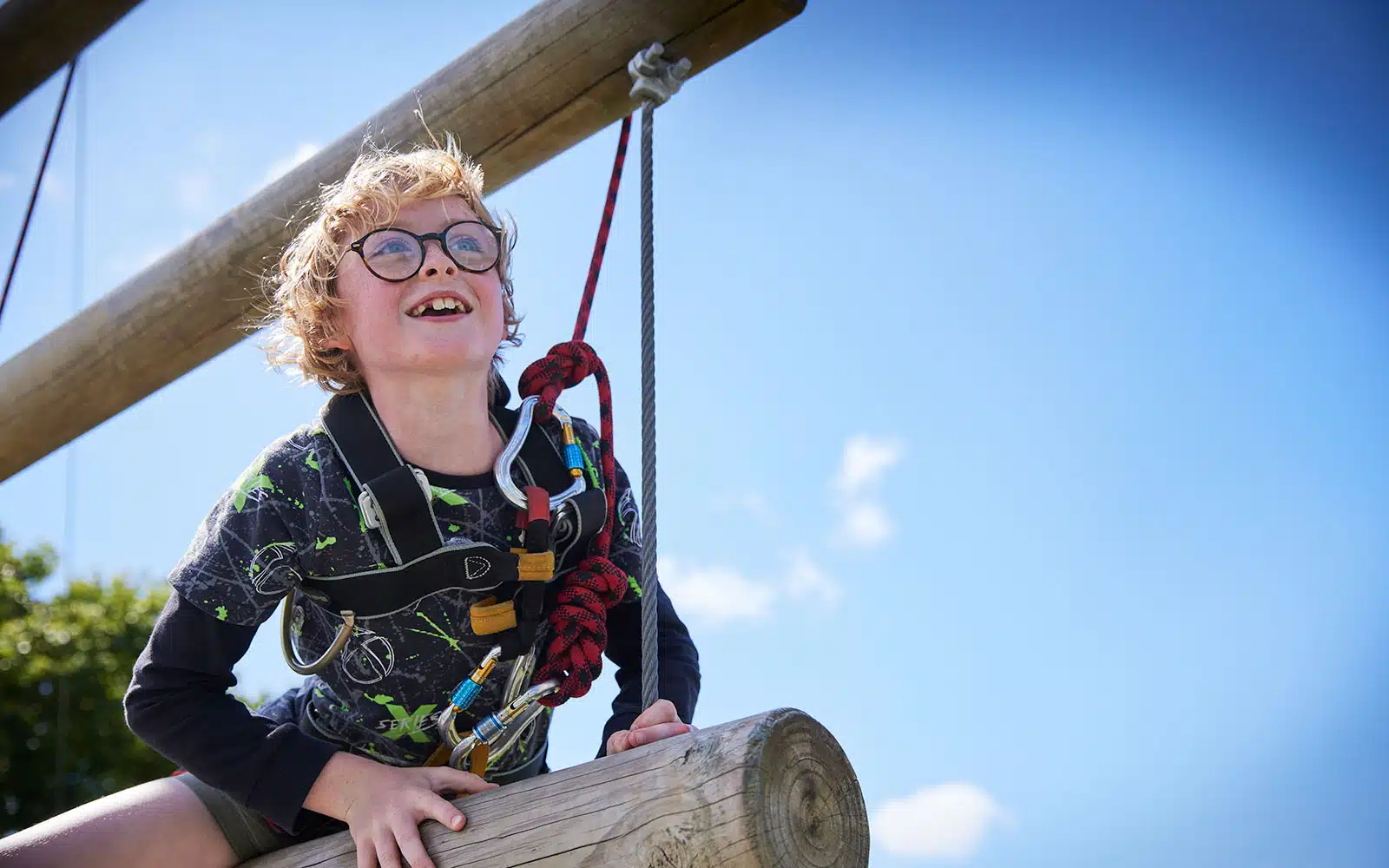 A young boy with glasses smiles while sitting atop a wooden play structure, holding onto a rope, on a sunny day at PGL Adventure Holidays.