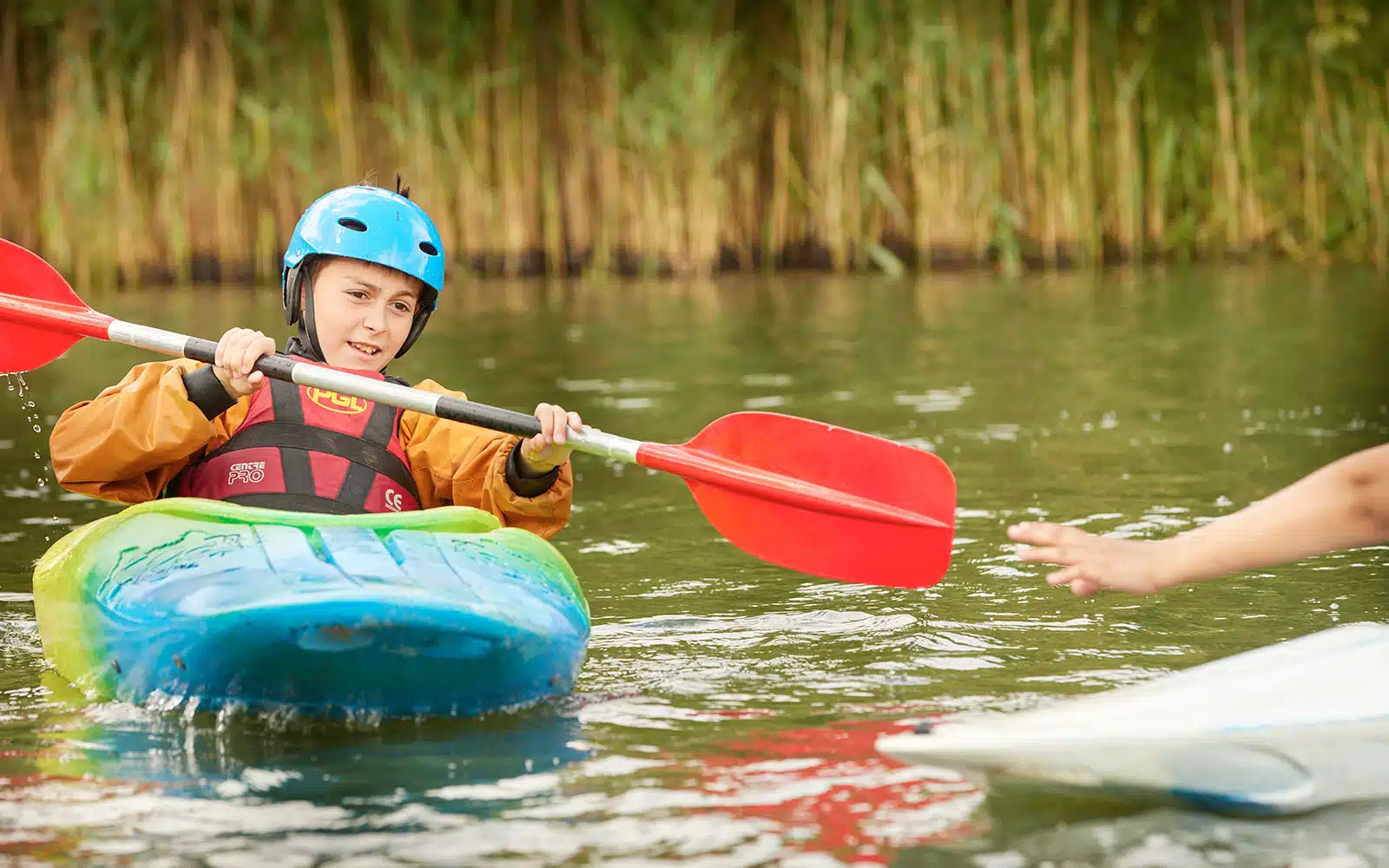 A young person in a blue helmet and orange jacket learning to kayak with PGL Adventure Holidays, receiving a paddle from another person's outstretched hand.