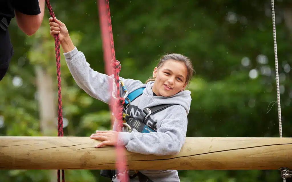 A girl in a harness smiles while participating in a PGL Adventure Holidays rope-based obstacle course outdoors, gripping a wooden beam with red ropes.