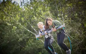 Two girls smiling and embracing while zip-lining against a backdrop of lush green trees during their PGL Adventure Holidays.