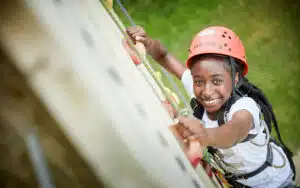 A young Black girl in a helmet and harness smiles while climbing on an outdoor PGL Adventure Holidays rope course.