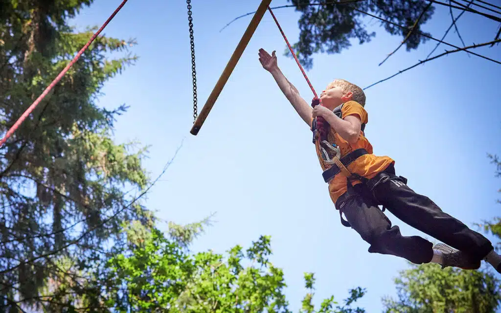 Young boy swinging on a rope at PGL Adventure Holidays, surrounded by trees on a sunny day.