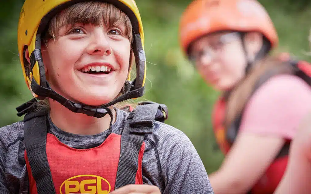 A smiling boy wearing a helmet and harness, with a girl in similar PGL Adventure Holidays gear slightly out of focus in the background outdoors.