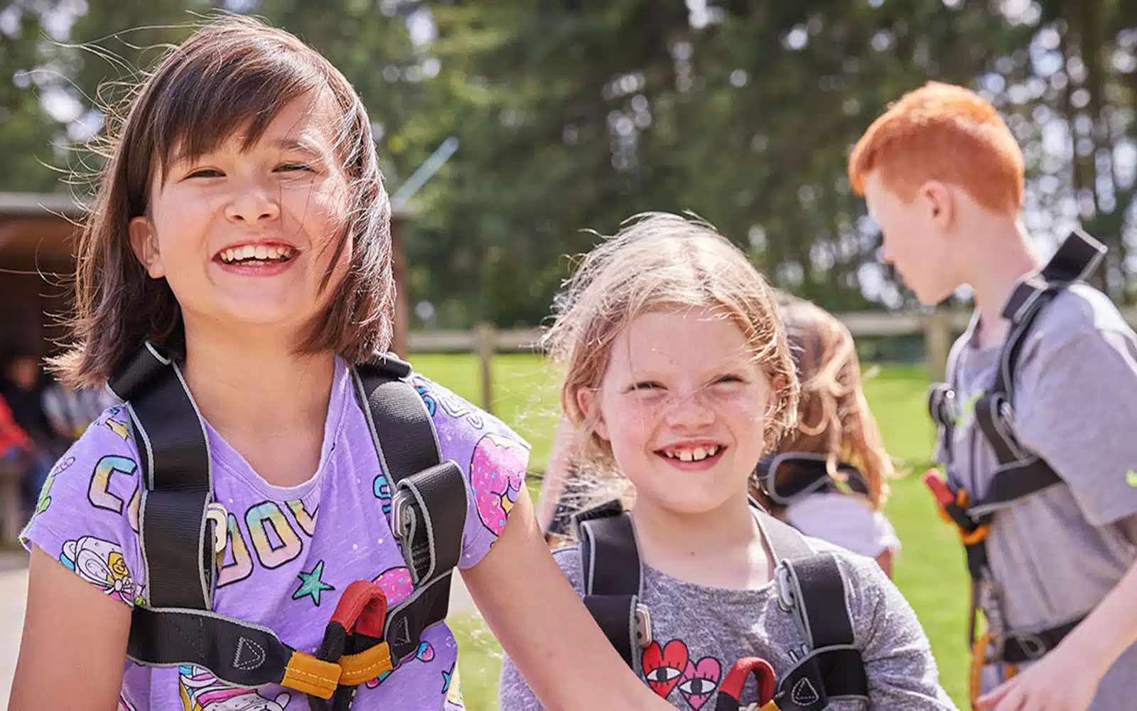 Two children smiling with harnesses on a sunny day at a PGL Adventure Holidays park, another child visible in the background.