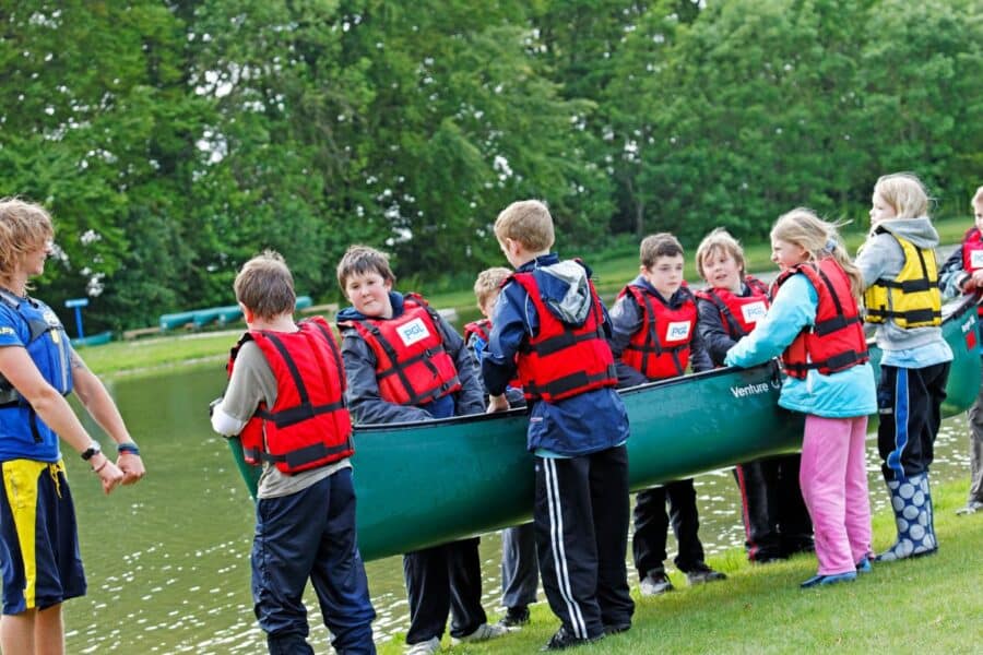 A group of children in life jackets receiving canoeing instructions from an instructor beside a lake.