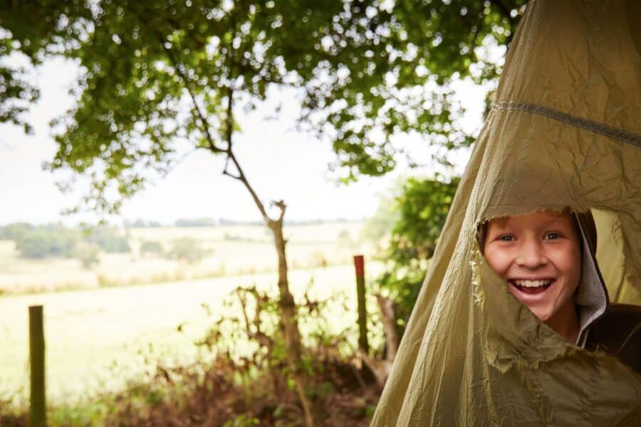 A child smiles while peeking out from a tent in a countryside setting with trees and a field in the background.