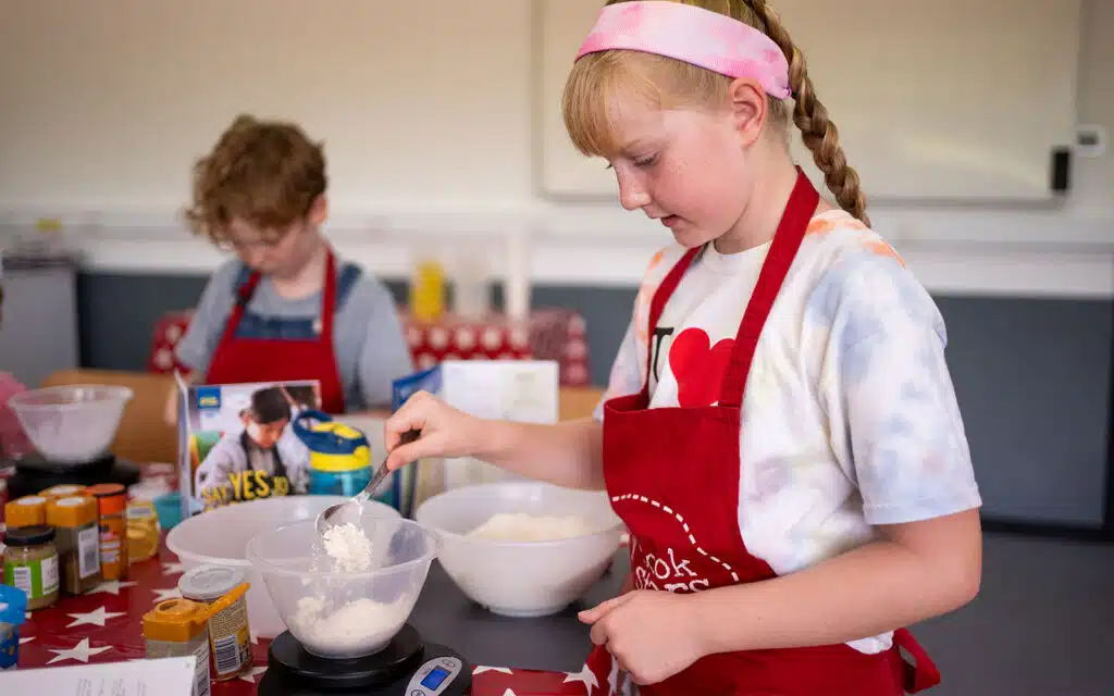 Young girl in a red apron and pink headband measuring flour with another child in the background also cooking.