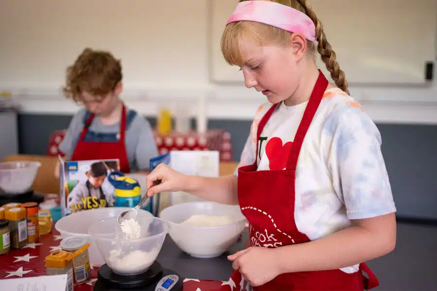 Young girl in a red apron and pink headband measuring flour with another child in the background also cooking.