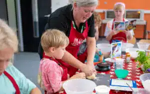 A cooking instructor helps a young boy knead dough during a children's cooking class, with other kids engaged in activities around them.
