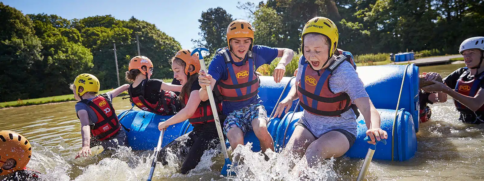 A group of people wearing helmets and life jackets paddle on a raft made of blue barrels in a sunny outdoor setting with trees and a pond. Water splashes around them as they navigate.