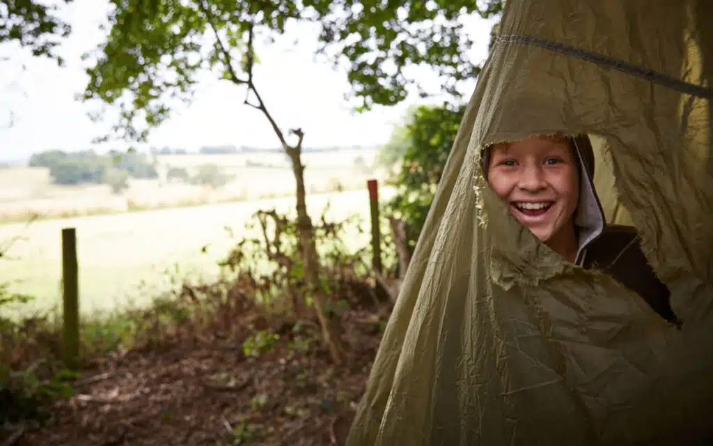 A cheerful young boy peeks out from a tent flap in a lush green camping area with a scenic countryside view in the background.