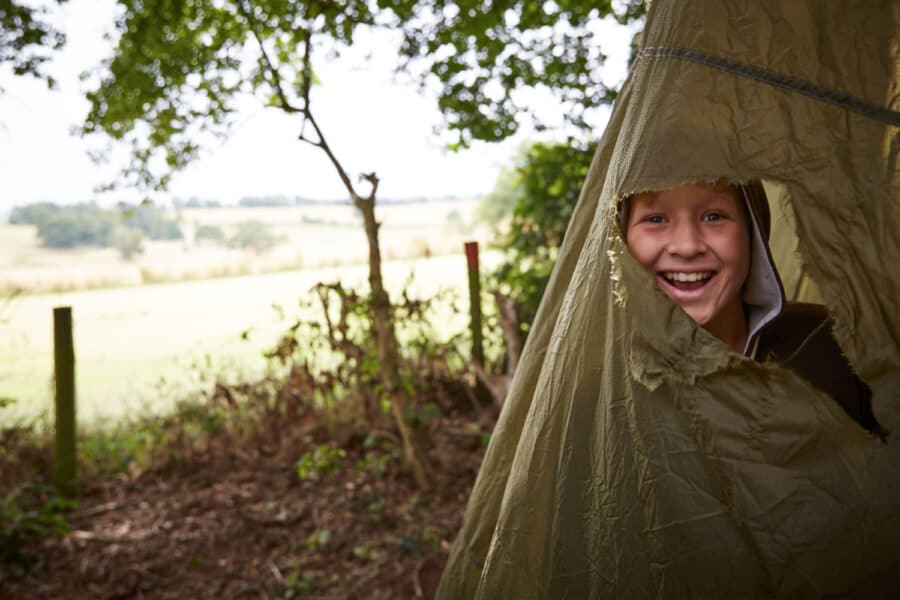 A cheerful young boy peeks out from a tent flap in a lush green camping area with a scenic countryside view in the background.