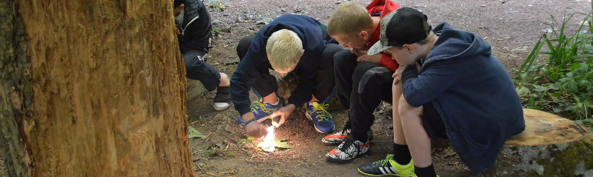 Four boys in a forest learning how to start a fire, gathered around small flames on the ground, with one actively lighting the fire.