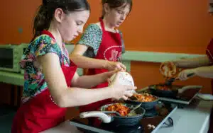 Two young girls in red aprons cooking in a kitchen during their PGL Adventure Holidays, transferring vegetables from a bowl to a skillet.