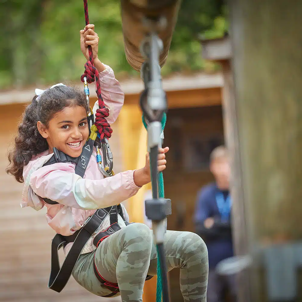A young girl with curly hair smiles while climbing ropes in an outdoor adventure park, wearing a safety harness and camouflage leggings.