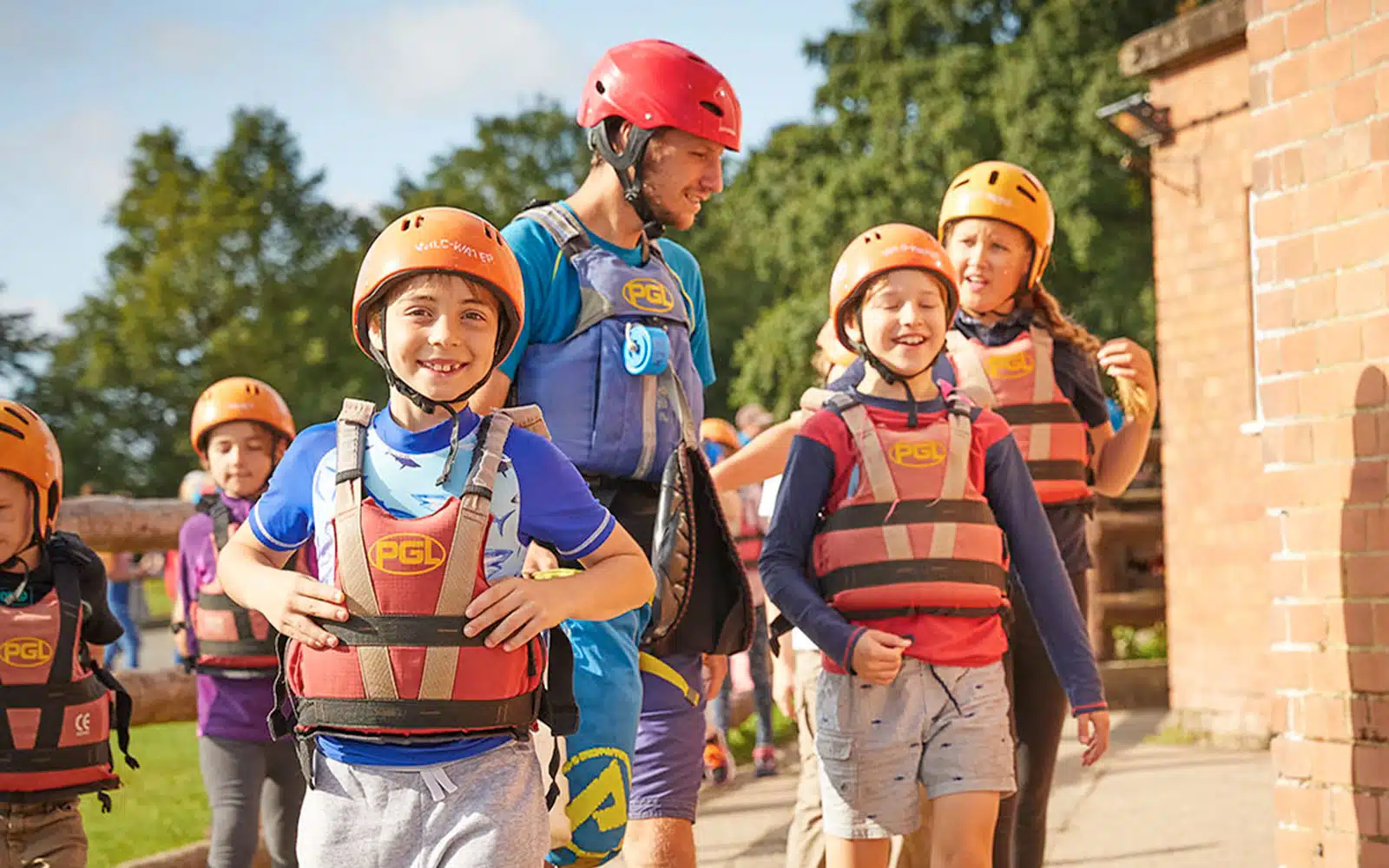 Group of children and an instructor from PGL Adventure Holidays, wearing safety helmets and life jackets, smiling and walking outdoors on a sunny day.