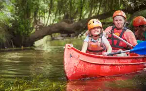Two people wearing helmets and life jackets paddling a red canoe through a tree-lined waterway on a PGL Adventure Holiday.