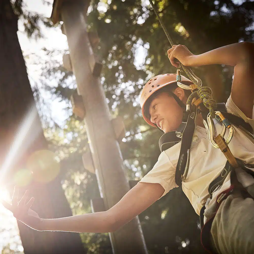 Person wearing a helmet and harness engages in a high ropes course amid tall trees, with sunlight filtering through the branches.