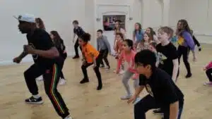 A diverse group of children at a PGL Adventure Holidays camp following a dance instructor in a spacious studio, mimicking his stance with focus and enthusiasm.