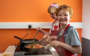 Two children cooking at a kitchen stove, both wearing aprons. The one in front is smiling while stirring food in a frying pan. An orange wall is in the background.