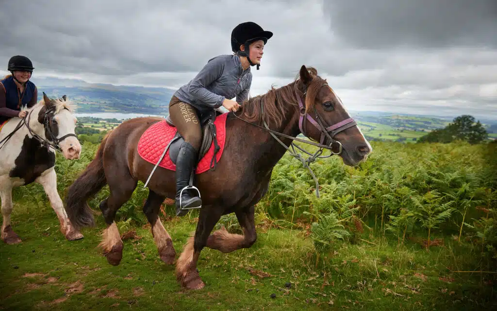 Two riders on horses trotting through a lush, hilly landscape under an overcast sky.