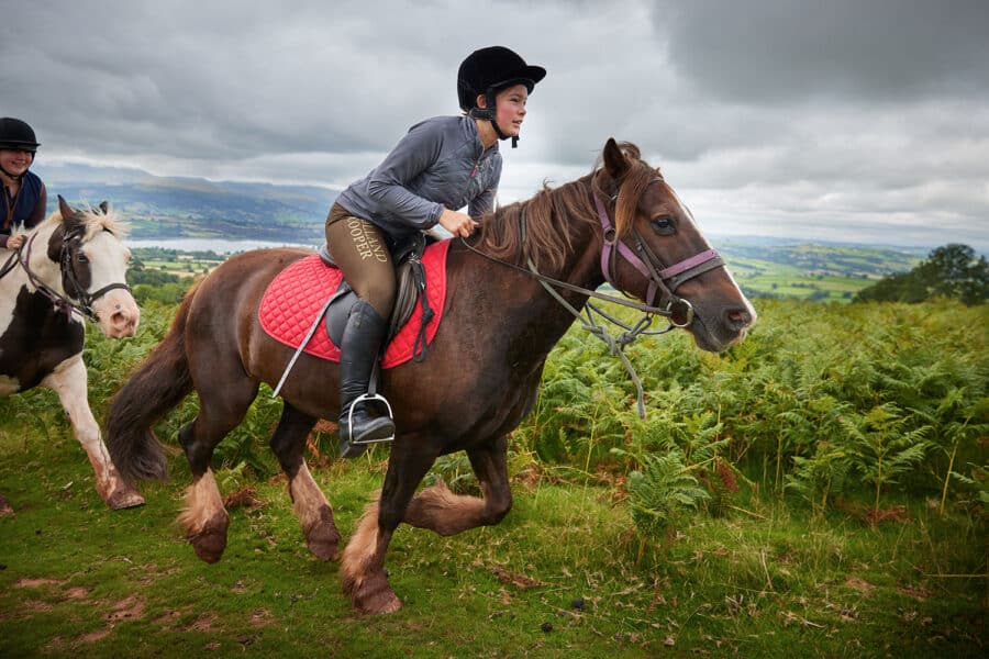Two riders on horses trotting through a lush, hilly landscape under an overcast sky.