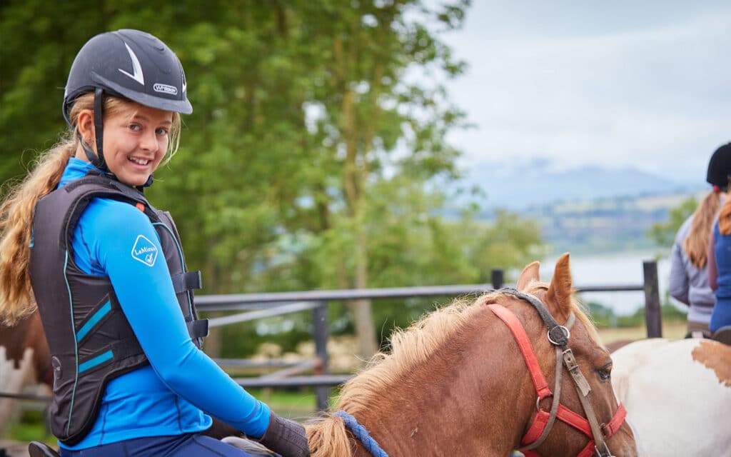 A smiling young woman wearing a helmet and body protector riding a chestnut horse in a fenced area with a scenic backdrop.