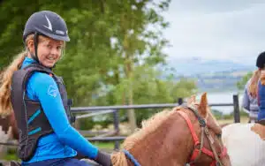 A smiling young woman wearing a helmet and body protector riding a chestnut horse in a fenced area with a scenic backdrop.
