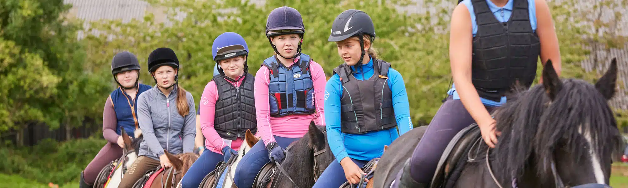 A group of young equestrians, wearing helmets and vests, riding horses outdoors, with a focus on one girl looking towards her left.