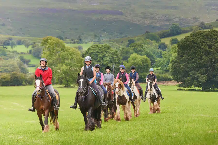 Group of seven people Pony Camp Trekker horseback riding across green field with hills in the background. All riders wearing helmets at Tregoyd House - Jul 27, 2024.