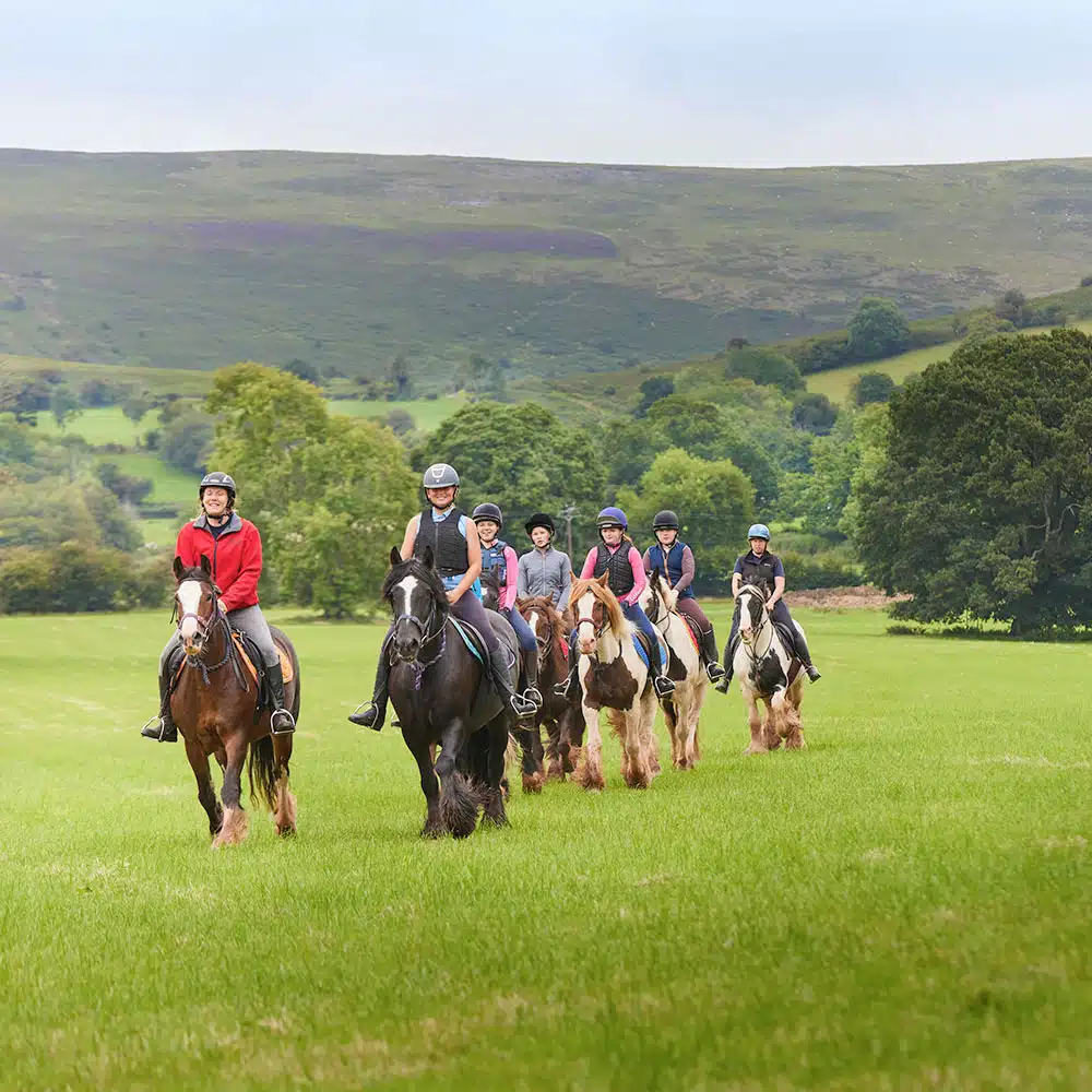 Group of seven people Pony Camp Trekker horseback riding across green field with hills in the background. All riders wearing helmets at Tregoyd House - Jul 27, 2024.