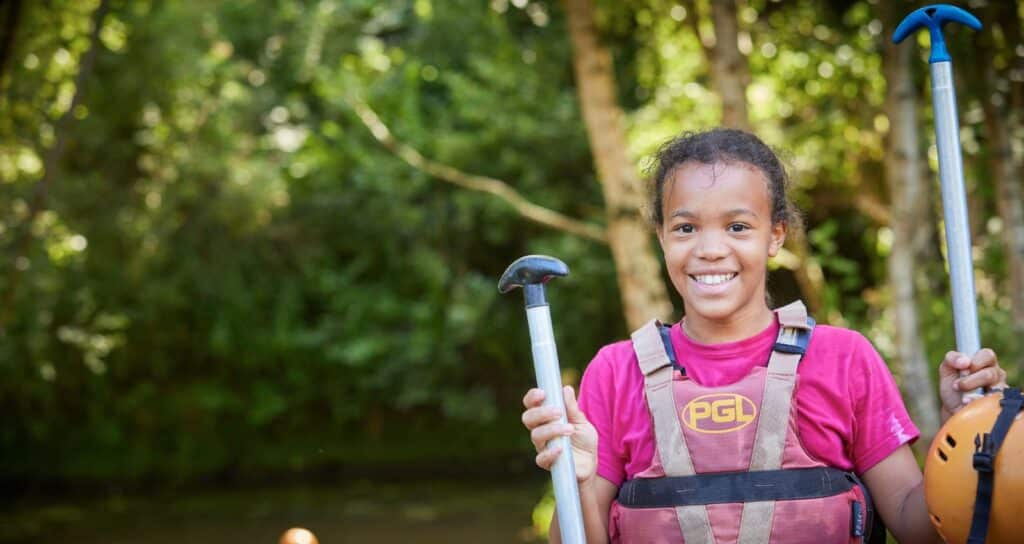 A young girl wearing a pink shirt and a safety vest holds paddles outdoors, smiling, with a forested background.