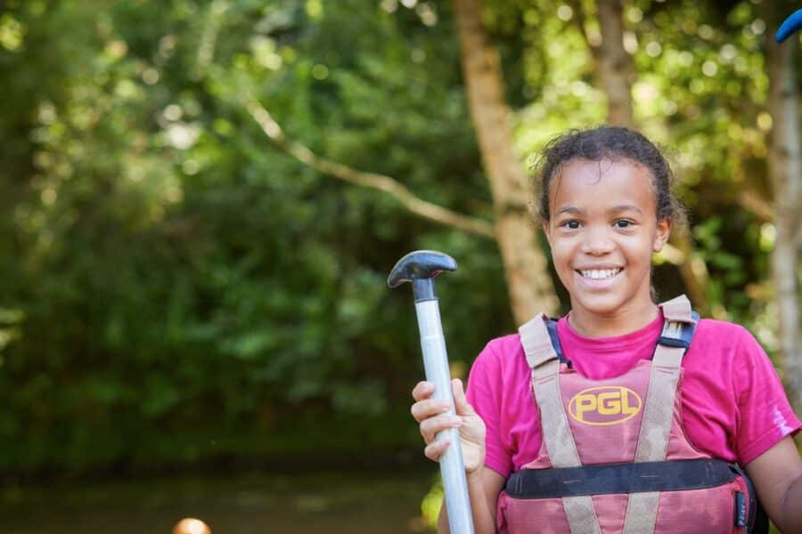 A young girl wearing a pink shirt and a safety vest holds paddles outdoors, smiling, with a forested background.