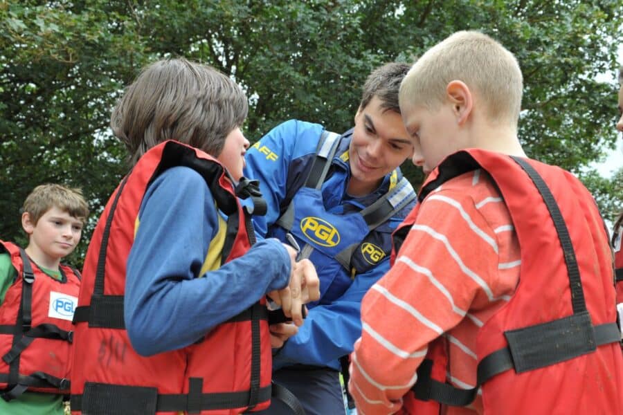 A group of children wearing life jackets stand close to an instructor who is helping one of them as others watch. Trees are visible in the background.