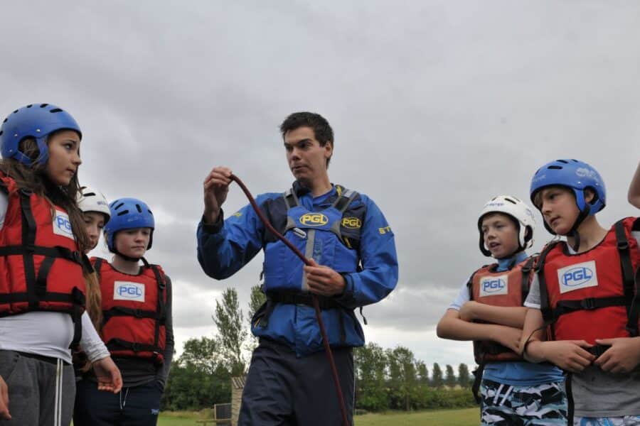 A man wearing a blue jacket demonstrates a rope-knot to a group of children in helmets and life vests standing outside on a cloudy day at an Outdoor Adventure Camp.
