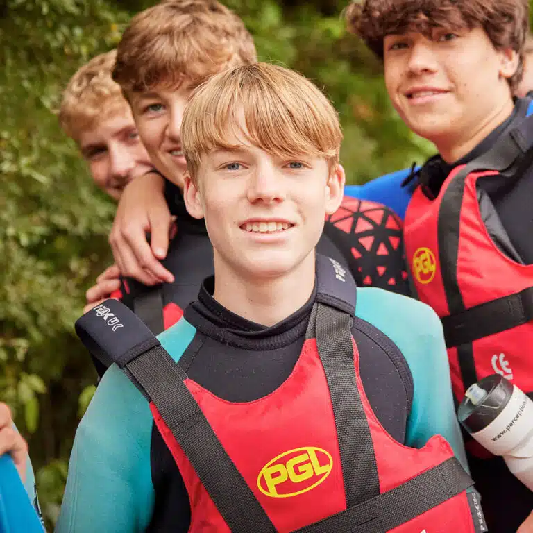 Four boys wearing red and black PGL life vests and wetsuits stand close together, smiling at the camera. Trees and greenery are visible in the background, embedding the joy of PGL Adventure Holidays.