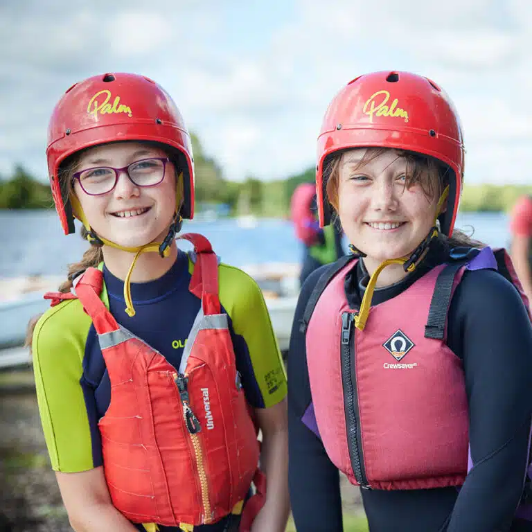 Two children wearing red helmets and life jackets smile at the camera, standing outdoors with a body of water and blurred background. Their excitement shines through as they enjoy their PGL Adventure Holidays experience.