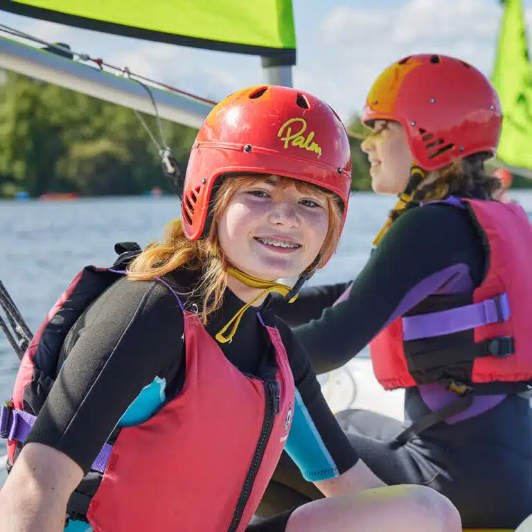 Two people wearing red helmets, life vests, and wetsuits sit on a boat with green sails. The person in the foreground is smiling at the camera. They are on a body of water with trees in the background, enjoying their PGL Adventure Holidays experience.