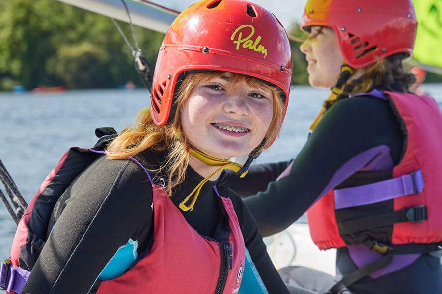 Two people wearing red helmets, life vests, and wetsuits sit on a boat with green sails. The person in the foreground is smiling at the camera. They are on a body of water with trees in the background, enjoying their PGL Adventure Holidays experience.