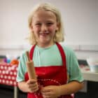 A child with blonde hair, wearing a red apron, holds a small rolling pin in a kitchen setting. A table with various cooking items is visible in the background, making it look like a scene from PGL Adventure Holidays.