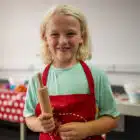 A child with blonde hair, wearing a red apron, holds a small rolling pin in a kitchen setting. A table with various cooking items is visible in the background, making it look like a scene from PGL Adventure Holidays.