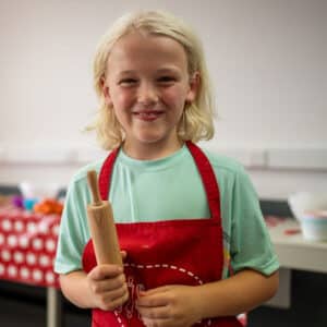 A child with blonde hair, wearing a red apron, holds a small rolling pin in a kitchen setting. A table with various cooking items is visible in the background, making it look like a scene from PGL Adventure Holidays.