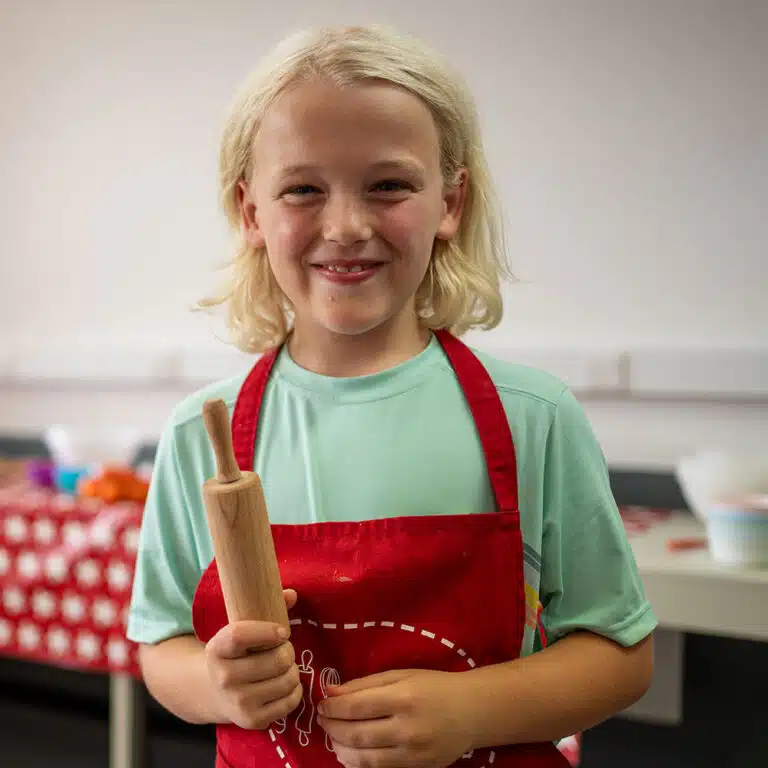 A child with blonde hair, wearing a red apron, holds a small rolling pin in a kitchen setting. A table with various cooking items is visible in the background, making it look like a scene from PGL Adventure Holidays.