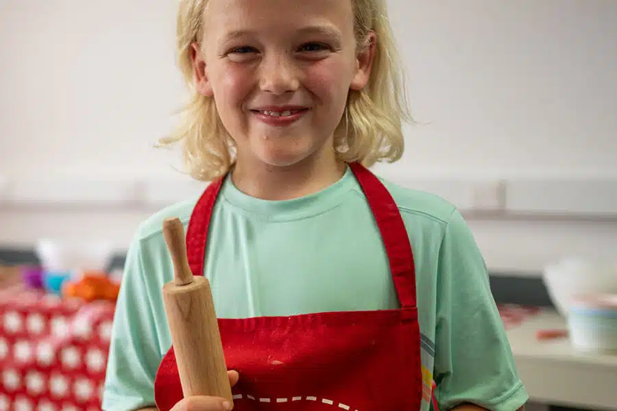 A child with blonde hair, wearing a red apron, holds a small rolling pin in a kitchen setting. A table with various cooking items is visible in the background, making it look like a scene from PGL Adventure Holidays.