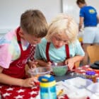 Two children in aprons are looking into a bowl with green ingredients on a red tablecloth, reminiscent of the fun-filled cooking sessions at PGL Adventure Holidays, while another person stands in the background.