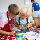 Two children in aprons are looking into a bowl with green ingredients on a red tablecloth, reminiscent of the fun-filled cooking sessions at PGL Adventure Holidays, while another person stands in the background.