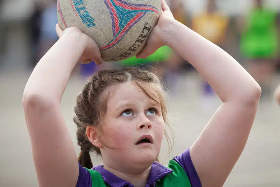 Young girl in a green and purple sports uniform, preparing to shoot a netball outdoors during her PGL Adventure Holiday. She is looking upwards, holding the ball above her head with both hands.